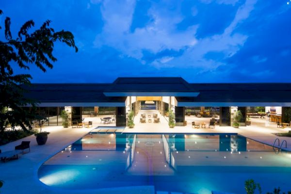 An illuminated modern building at dusk features a large, inviting swimming pool in the foreground and open seating areas inside.
