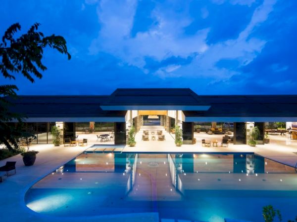 An illuminated modern building at dusk features a large, inviting swimming pool in the foreground and open seating areas inside.