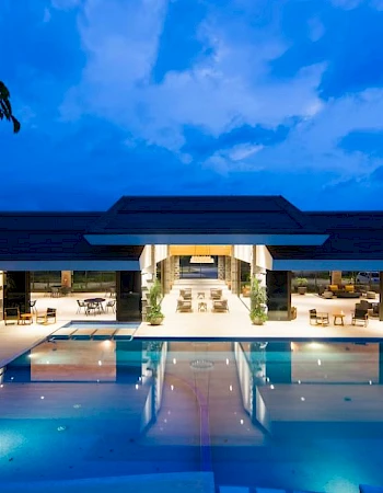The image shows a luxurious resort with a large illuminated pool, lounge chairs, and a modern open-air building illuminated at twilight, ending the sentence.