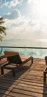 A serene poolside scene overlooking the ocean, with lounge chairs and umbrellas on a wooden deck under a bright, partly cloudy sky.