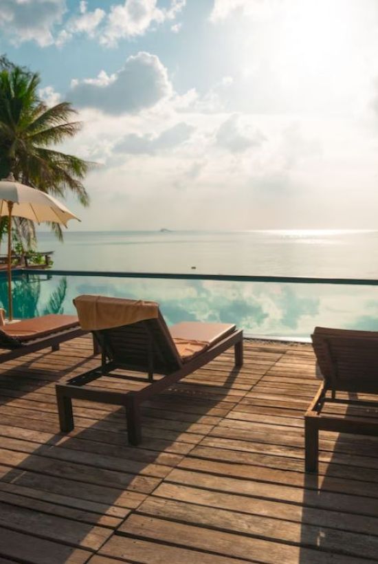 A serene poolside scene with lounge chairs, umbrellas, palm trees, a wooden deck, and a stunning view of the sea under a bright sky.