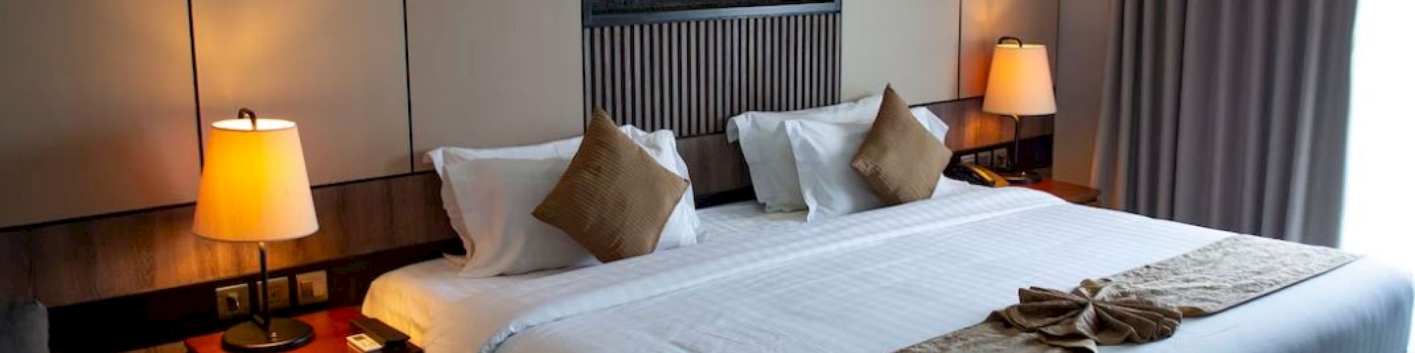 The image shows a hotel room with a neatly made king-sized bed, nightstands with lamps, a wall-mounted decor piece, and curtains.