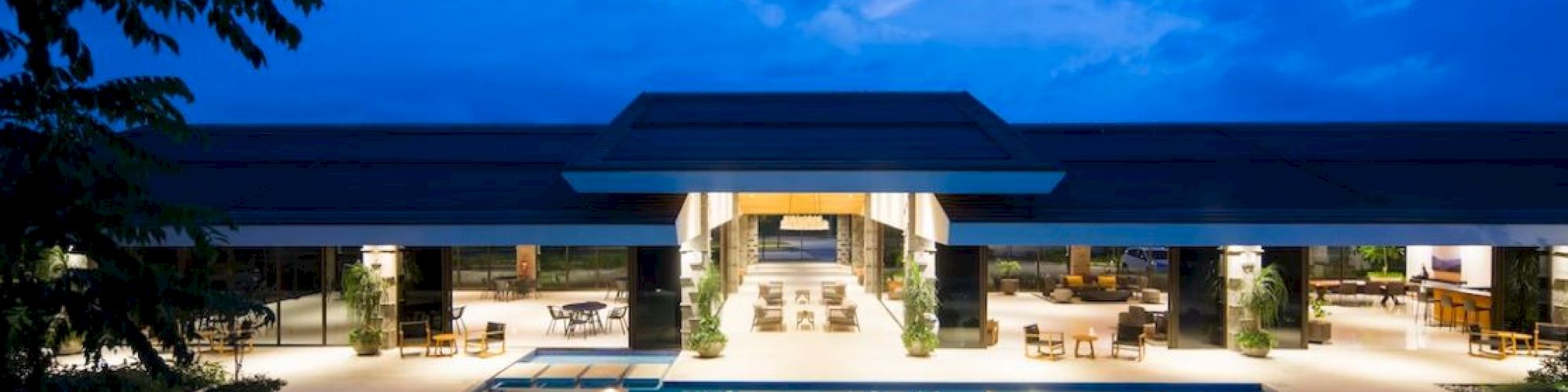 A luxurious, well-lit building with an expansive swimming pool under a twilight sky, surrounded by lounge chairs and greenery.