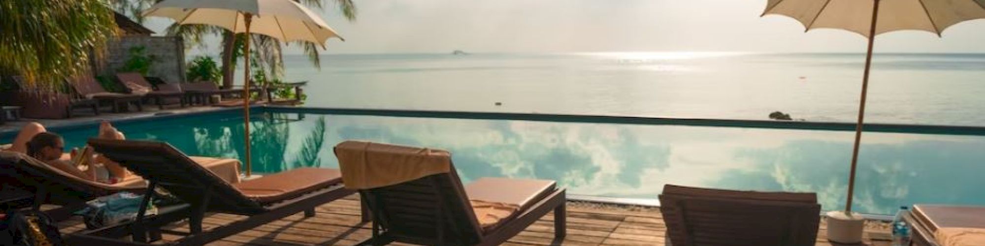 The image shows lounge chairs and umbrellas on a wooden deck by an infinity pool, overlooking a calm sea with a bright sun in the sky.