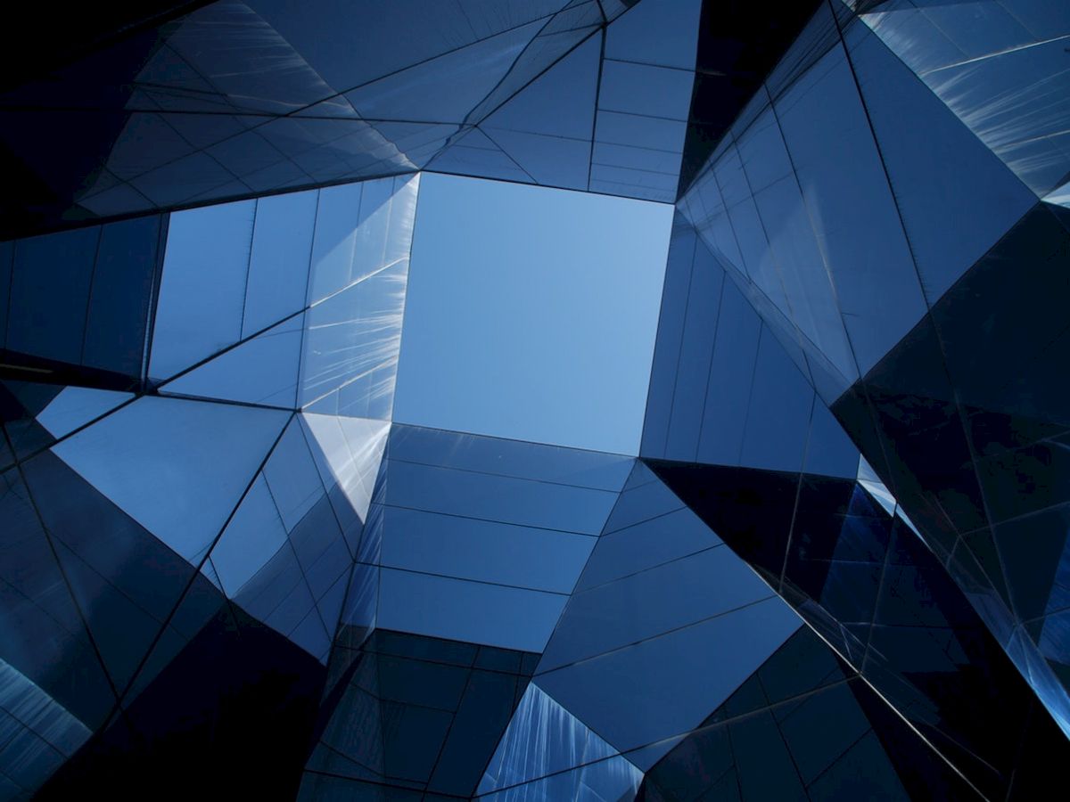 The image shows a modern architectural structure featuring reflective glass panels, with a view of the sky through a geometric opening in the center.