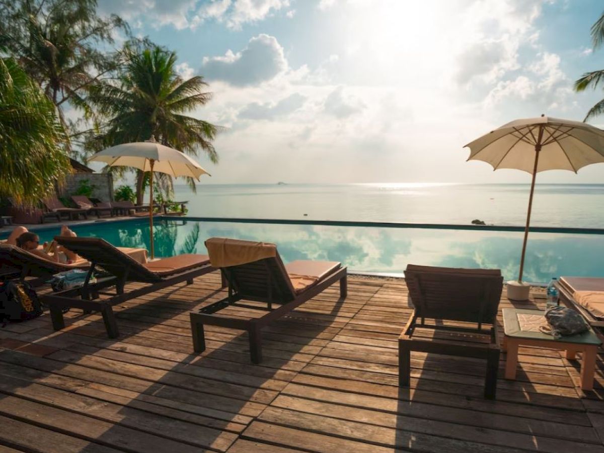 A serene poolside view with lounge chairs, umbrellas, palm trees, and a beautiful ocean horizon under a partly cloudy sky.