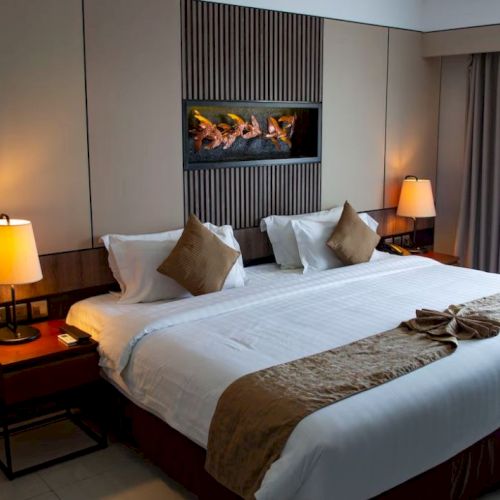 A well-decorated hotel room features a large bed with brown pillows, two bedside lamps, a modern fireplace, and a window covered by curtains.