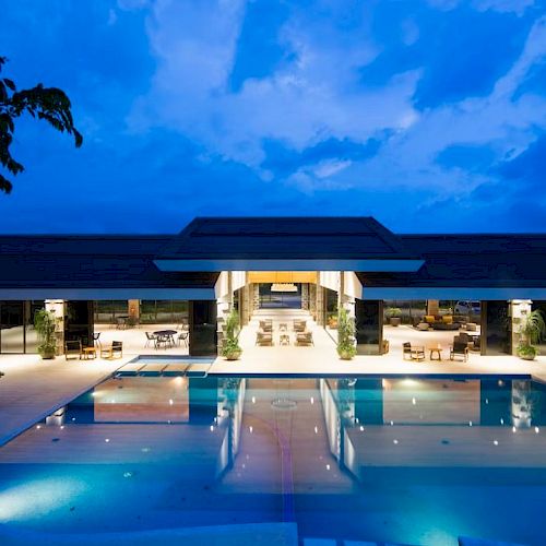 A luxurious open-air building with a large illuminated swimming pool in front, surrounded by lounge chairs and tables under a blue evening sky.