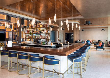 This image shows a modern bar with a wooden countertop, multiple bottles on shelves behind, stylish hanging lights, and navy blue bar stools.