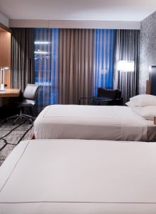 A modern hotel room with two neatly made beds, a wall-mounted TV, a desk and chair by the window, and stylish contemporary decor.