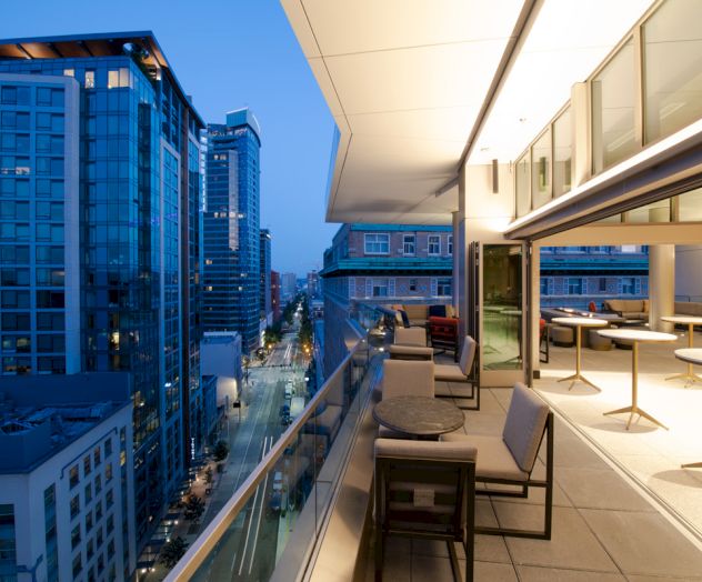 A cityscape featuring modern high-rise buildings at dusk, viewed from an elegant balcony with seating and tables, overlooking a well-lit street below.