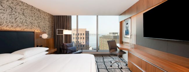 This image shows a modern hotel room with a large bed, a desk, a chair, a flat-screen TV, and a window with a city view in the background.