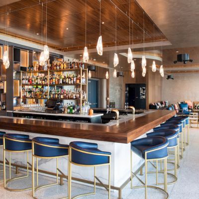This image features a modern bar with blue stools, a wooden countertop, hanging lights, and a fully stocked bar. Comfortable seating is visible in the background.