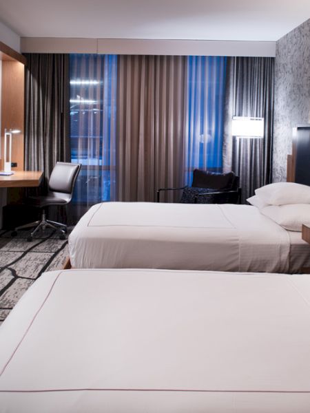 A hotel room with two beds, a large TV, a work desk, and modern decor featuring a patterned wall and floor-to-ceiling windows with curtains.