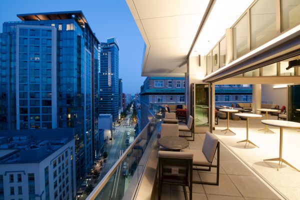 A modern urban balcony at dusk with seating and tables overlooks a city street, adjacent to a tall glass high-rise building.