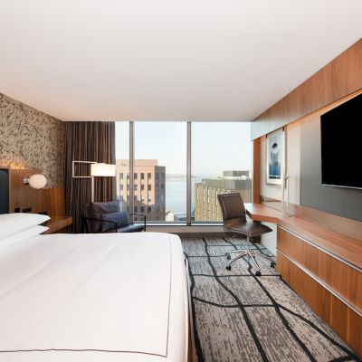 A modern hotel room with a large bed, desk, chair, and a wall-mounted TV. There is a window with a cityscape view in the background.