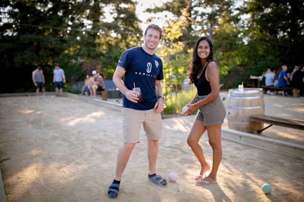 Two people having fun outdoors, holding bocce balls, with others sitting in the background near benches and tables on a sunny day.