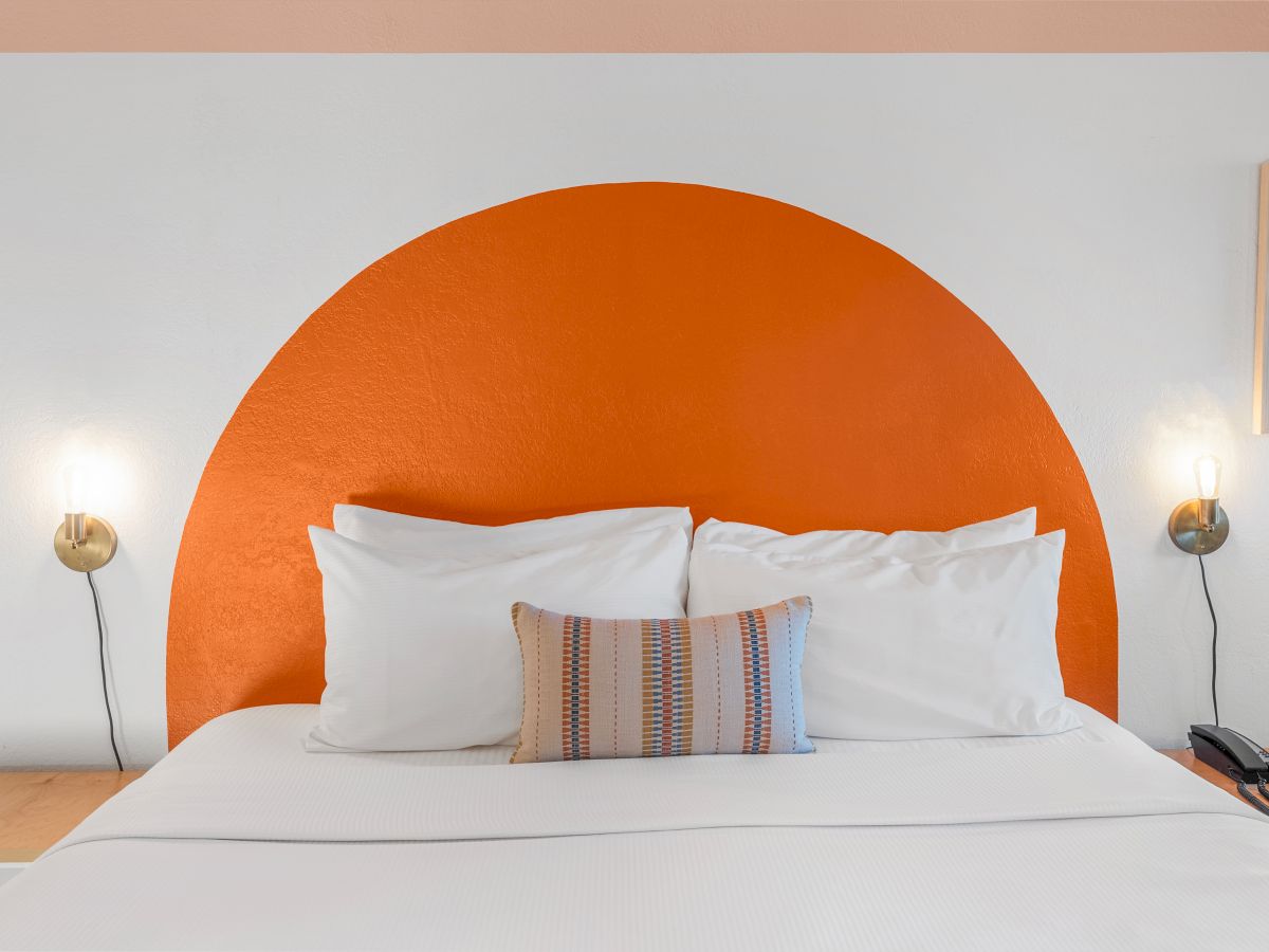 A neatly made bed with white linens and a colorful cushion, flanked by nightstands and lamps, has an orange circular headboard wall decoration.