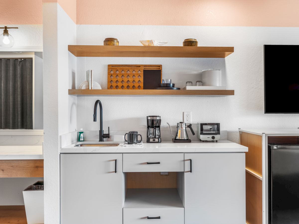 A small kitchenette with a sink, coffee maker, toaster, and shelves holding various kitchen items. A TV is mounted on the wall.