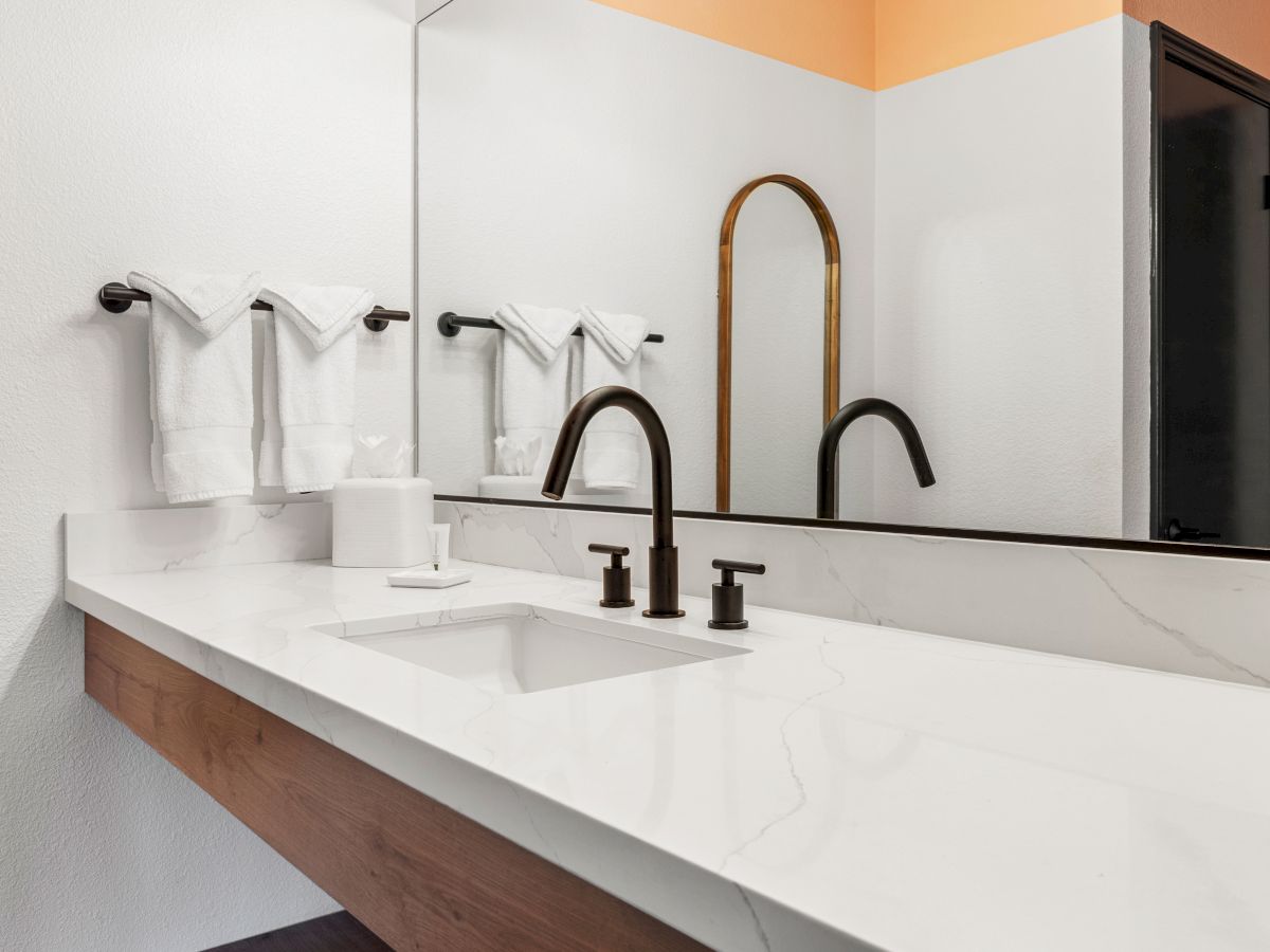 A modern bathroom with a white countertop, black faucet, large mirror, mounted towel rack holding white towels, and a gold arch mirror is shown.