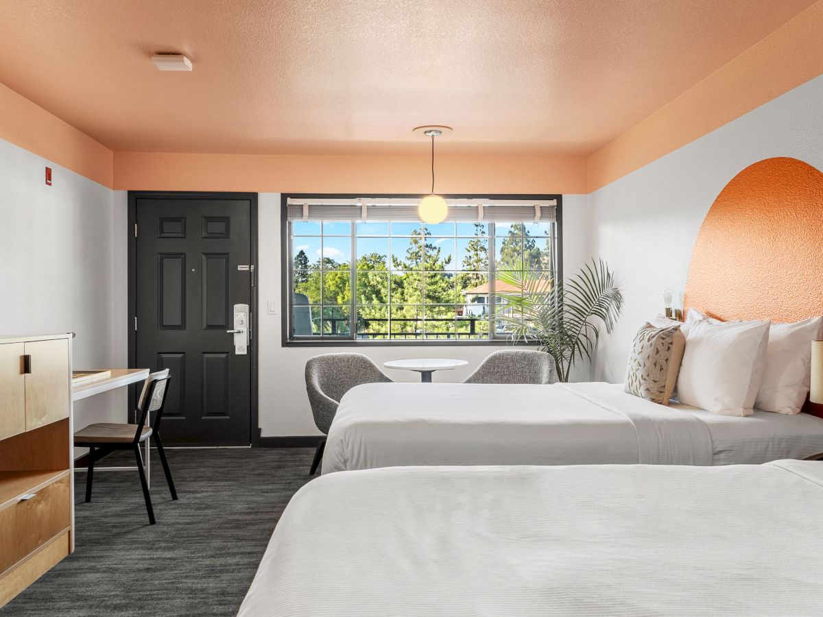 This image shows a modern hotel room with two beds, a desk with a chair, a window with a view, and an orange accent wall.