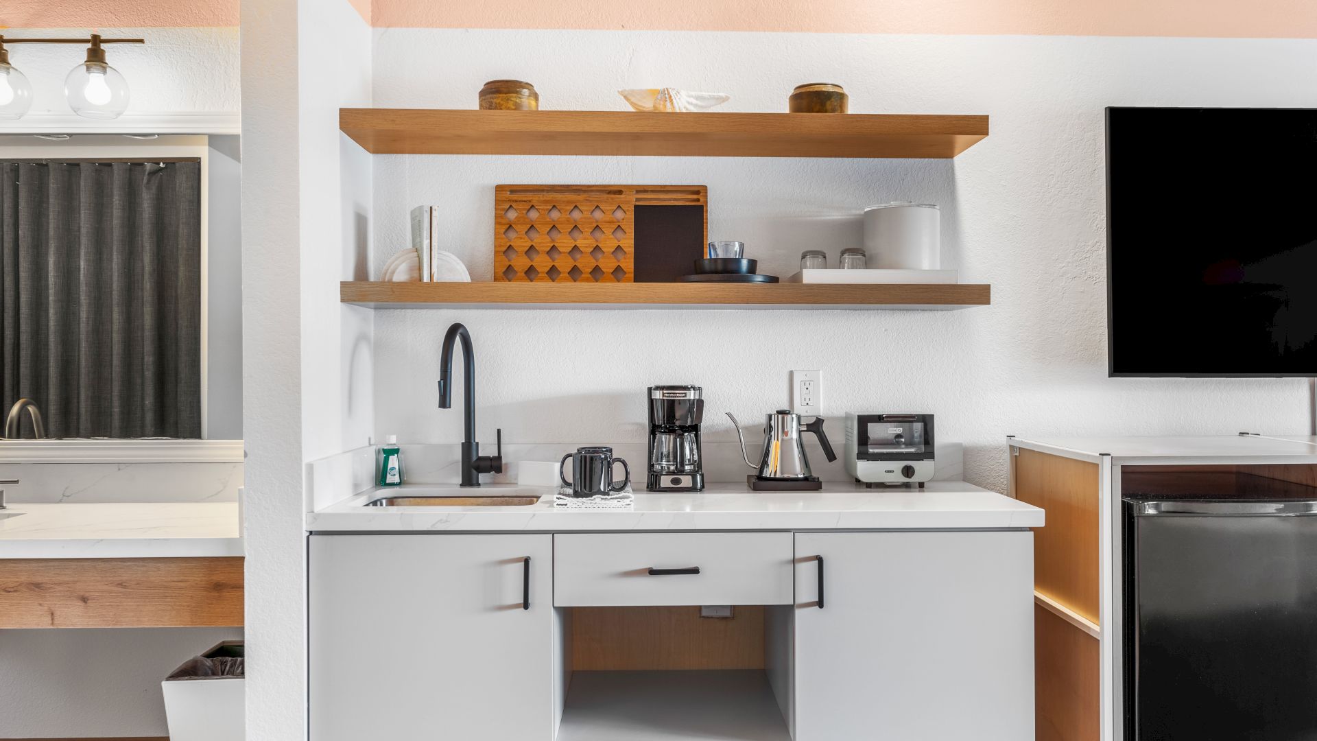 A modern kitchenette features a sink, coffee maker, toaster, shelves with d&eacute;cor, and appliances, alongside a bathroom area to the left and a TV to the right.
