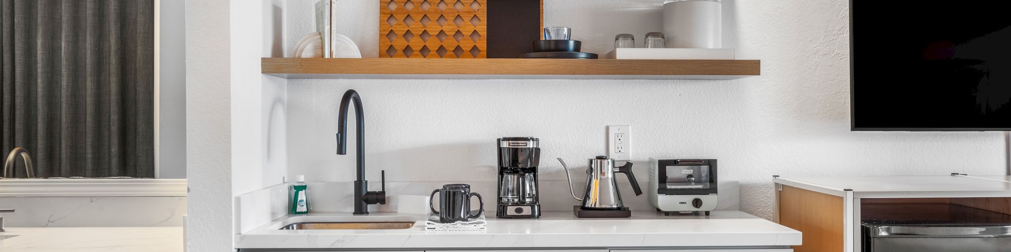 A modern kitchenette features a sink, coffee maker, toaster, shelves with décor, and appliances, alongside a bathroom area to the left and a TV to the right.