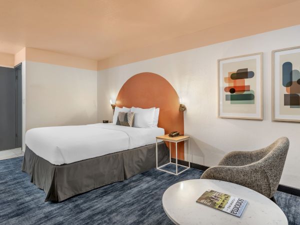 The image shows a modern hotel room with a bed, abstract art, a chair, a small table, and a magazine titled "Explore" on a round table.