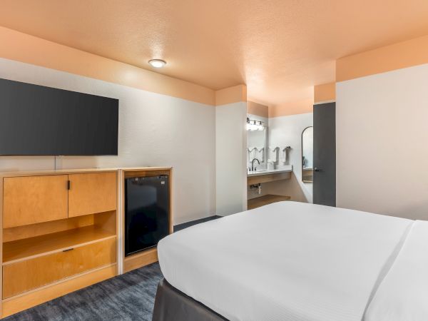 The image shows a hotel room with a bed, TV, wooden cabinet, mini fridge, and a bathroom area with a mirror and lights.