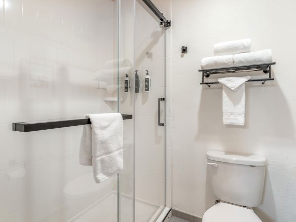 A bathroom with a glass shower, stocked towel shelf, toilet, and two dispensers mounted inside the shower area.