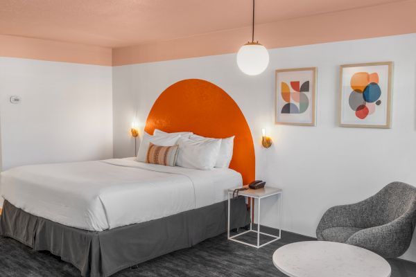 A modern hotel room with a bed, side table, armchair, and wall decor. A round light hangs from the ceiling, and orange accents add a pop of color.