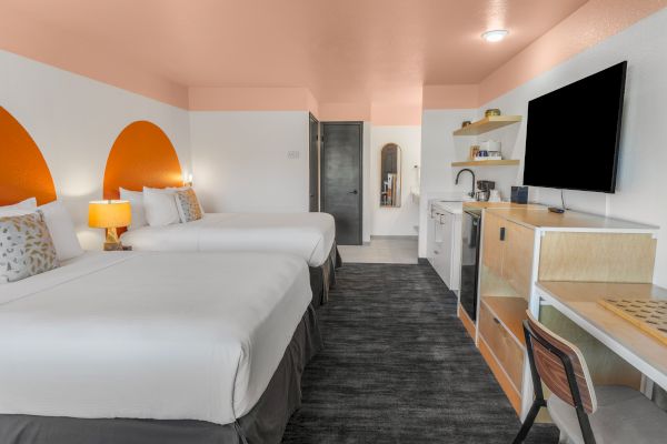 A modern hotel room with two beds, a wall-mounted TV, a desk, a mini-fridge, and orange accents on the walls and ceiling.