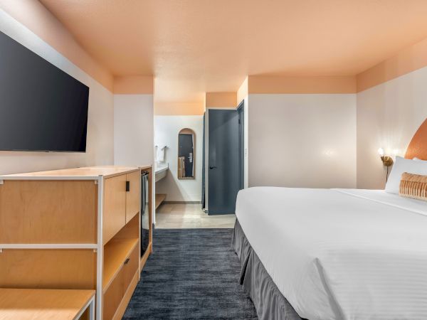 A modern hotel room with a king-sized bed, wall-mounted TV, light wood furniture, and a small fridge. Bright and minimalist design.