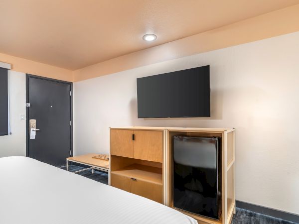 A hotel room featuring a bed, a flat-screen TV on the wall, a mini fridge, wooden cabinet, and a door with a sign hanging on the handle.