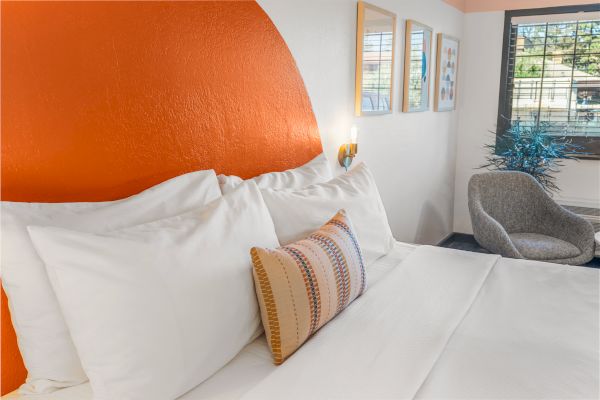 A stylish bedroom features a bed with white linens and a decorative pillow, an orange headboard, framed artwork, a plush chair, and a window.