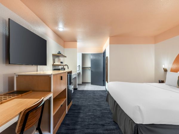 A modern hotel room with a bed, TV, desk, kitchenette, and a walk-in area leading to a bathroom.