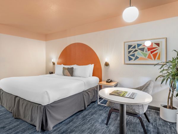A cozy hotel room with a large bed, modern decor, a geometric art piece, small table with a chair, and a plant in the corner.