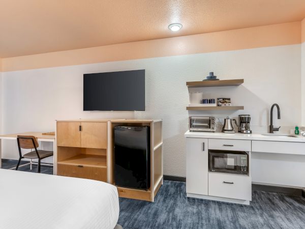 The image shows a hotel room with a TV, a desk with a chair, a mini-fridge, microwave, toaster oven, coffee maker, and a sink.
