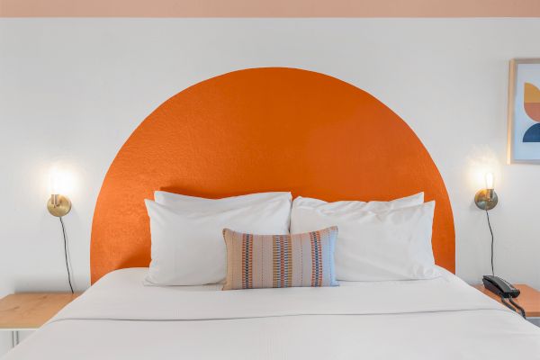 A neatly made bed with white linens, an orange semi-circular headboard, two bedside lamps, and a nightstand on each side.