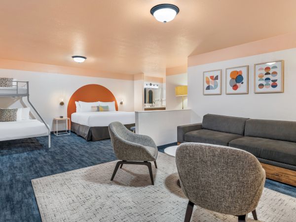 The image shows a modern hotel room with a double bed, bunk beds, seating area, and abstract art on the walls.