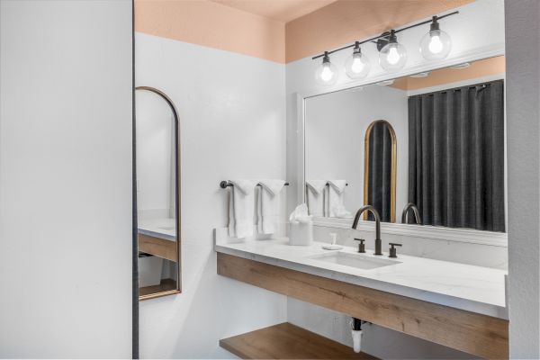 The image shows a modern bathroom with a large mirror, a sink with black fixtures, a light fixture with four bulbs, and white and wooden accents.