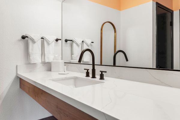 A modern bathroom vanity with a large mirror, black faucets, white countertop, and neatly folded towels hanging on a rack.