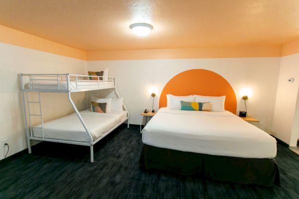 The image shows a hotel room with a queen-sized bed, a bunk bed, two wall-mounted reading lights, and a round orange design on the wall behind the queen bed.