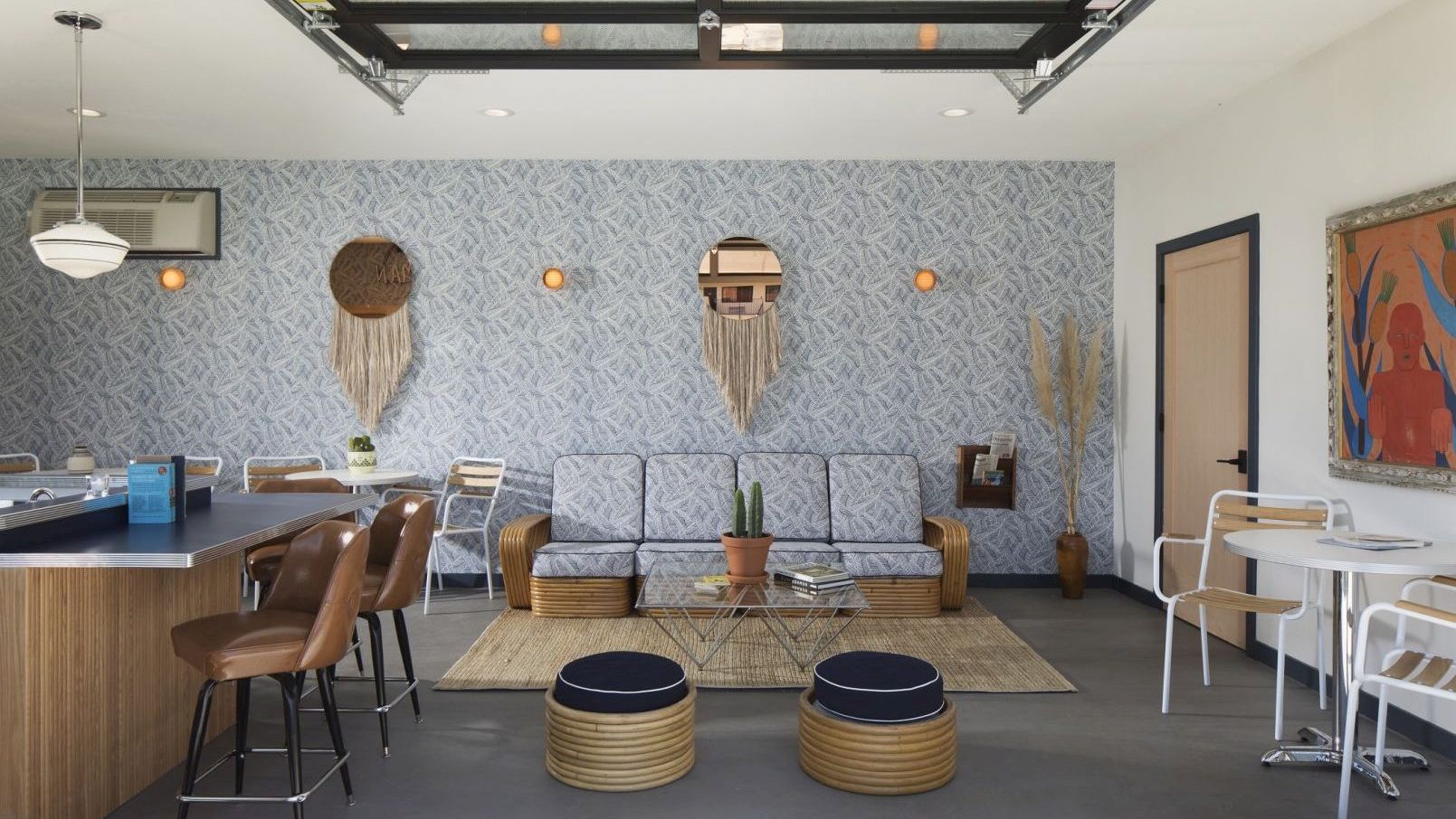 A modern lounge area with a sofa, poufs, wall art, and plants. There's a bar with stools on one side and small tables and chairs on the other side.