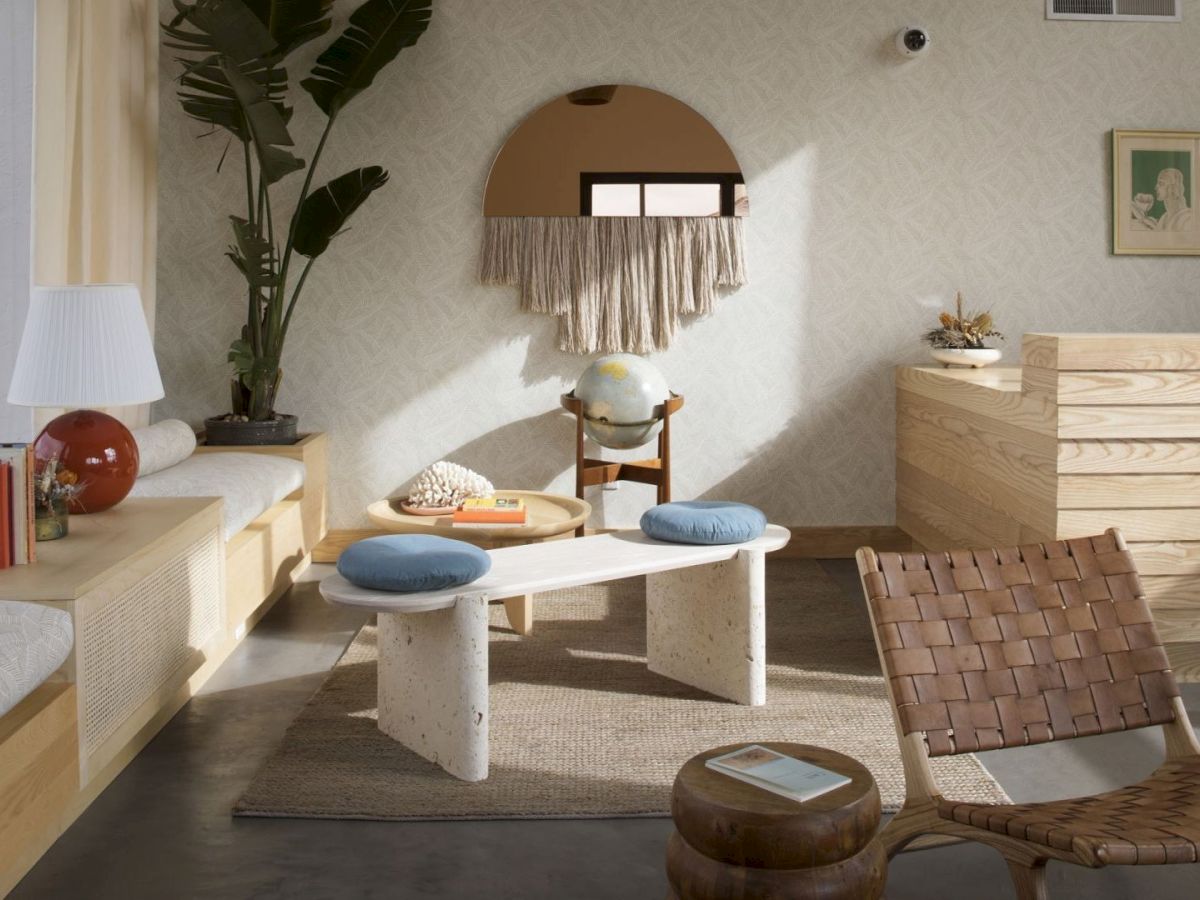 A cozy room with modern decor: wicker chair, globe on wooden side table, round mirror with fringe, plants, and a mix of wooden furniture.