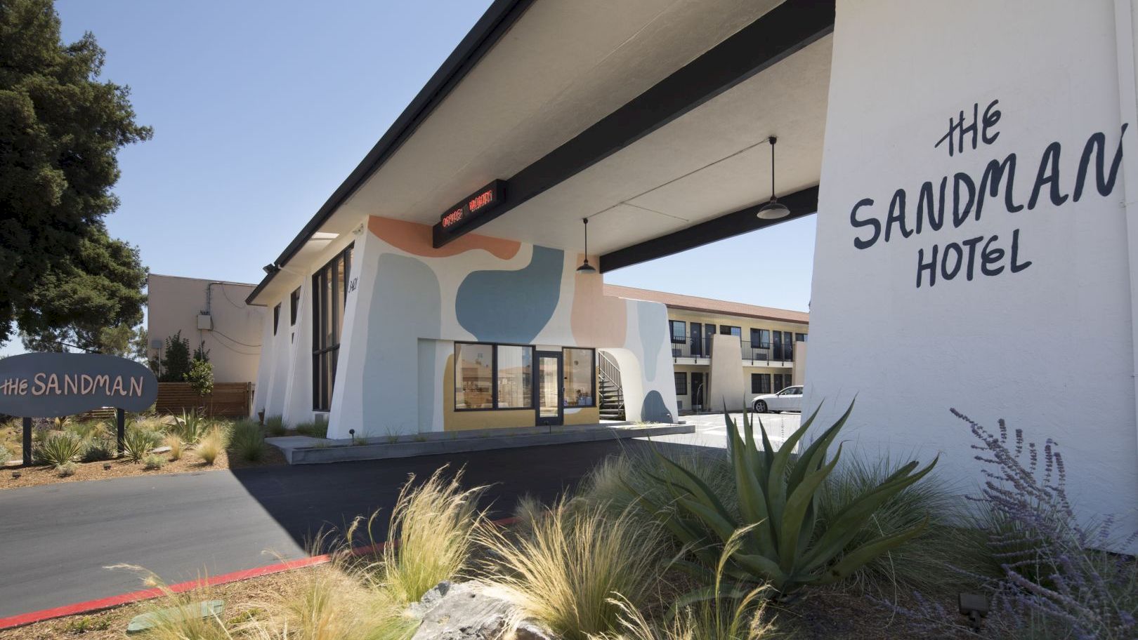 The image shows the exterior of The Sandman Hotel, featuring modern architecture, a welcoming entrance, and a landscaped area with plants.