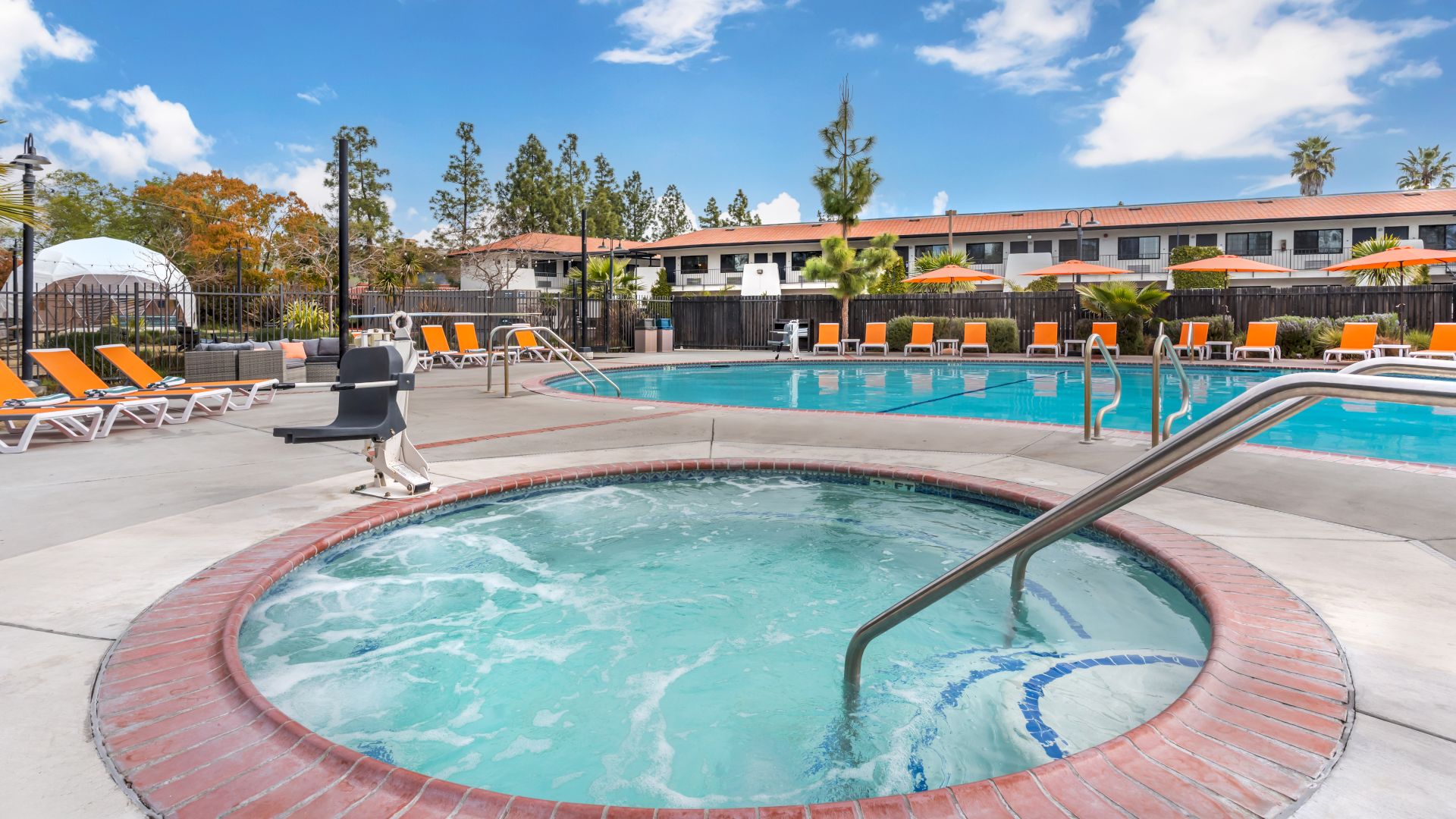 The image shows a swimming pool and hot tub area with lounge chairs and a building in the background under a partly cloudy sky.