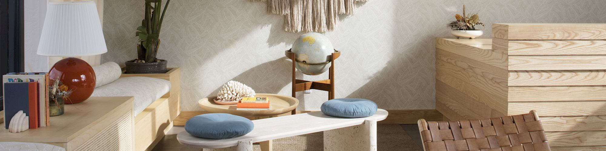 The image shows a cozy, modern living room with a wooden chair, cushions on a white table, potted plants, and a decorative wall mirror with fringe.