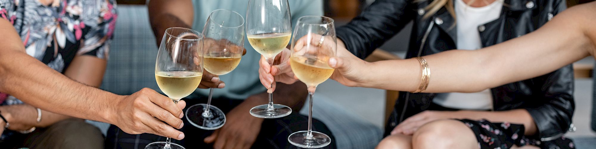 A group of friends is toasting with glasses of white wine at a table with food containers and a wine bottle in a relaxed setting.