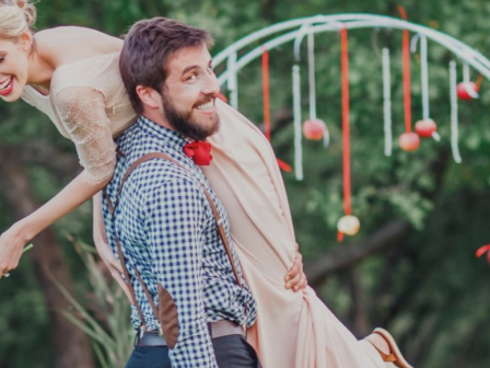 A man in suspenders carries a woman over his shoulder; they are smiling near an outdoor decorative arch with hanging ornaments in a greenery setting.